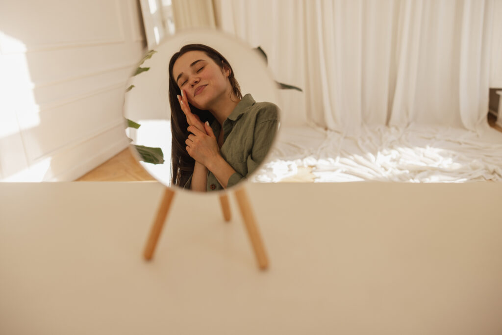 Your Personal Superhero: Finding Confidence and Self-Love Through Intimate Wellness 5 Woman looking at her reflection in mirror, touching her face with self-love and confidence