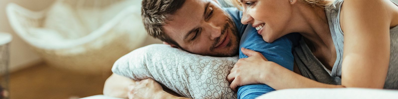 A couple lying close together in bed, talking and smiling in soft morning light