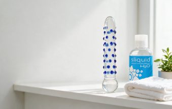 Clear textured glass dildo beside a bottle of water-based lubricant on a bathroom shelf